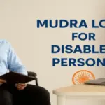 A confident Indian man in a wheelchair wearing a light blue shirt sits at a desk with a laptop, smiling while holding a portfolio. Text on the wall reads 