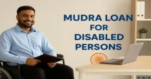 A confident Indian man in a wheelchair wearing a light blue shirt sits at a desk with a laptop, smiling while holding a portfolio. Text on the wall reads "Mudra Loan for Disabled Persons" alongside an Ashoka Chakra, symbolizing government support for physically disabled entrepreneurs in India. The background is a clean, modern office setting.